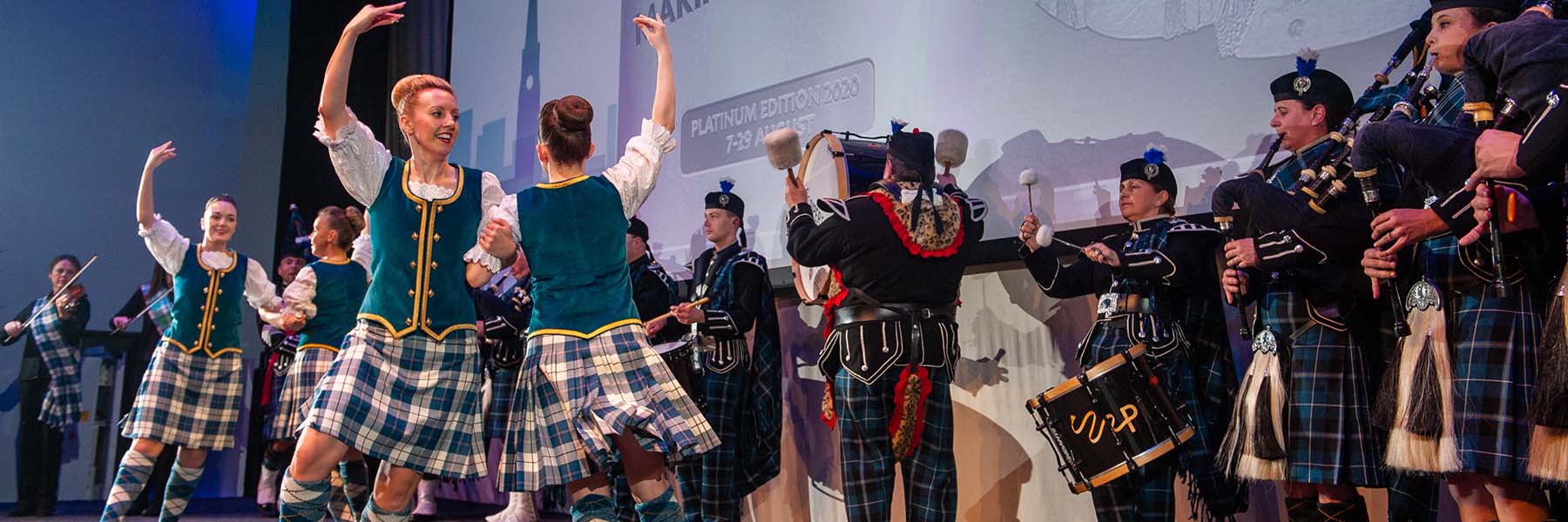 A stage of Scottish Highland dancers in traditional dress dancing to a live Pipe band
