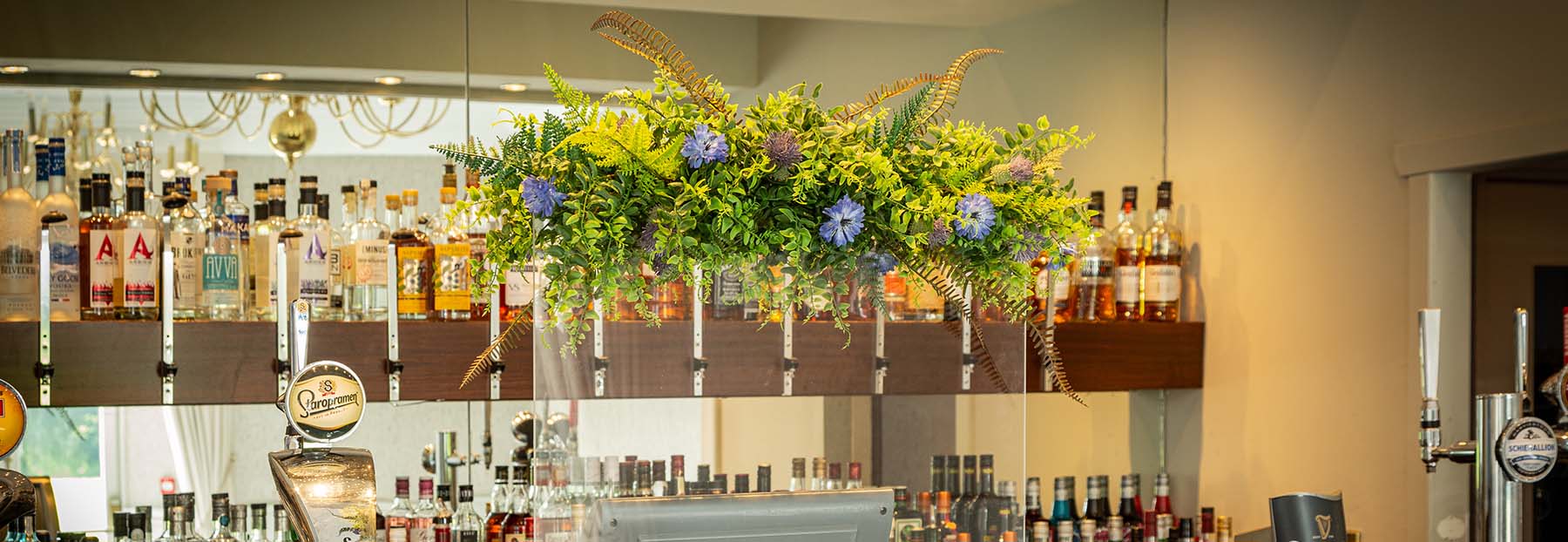 Hotel Bar with a PPE screen in front of the till topped with floral display of artificial foliage, flowers, and thistles