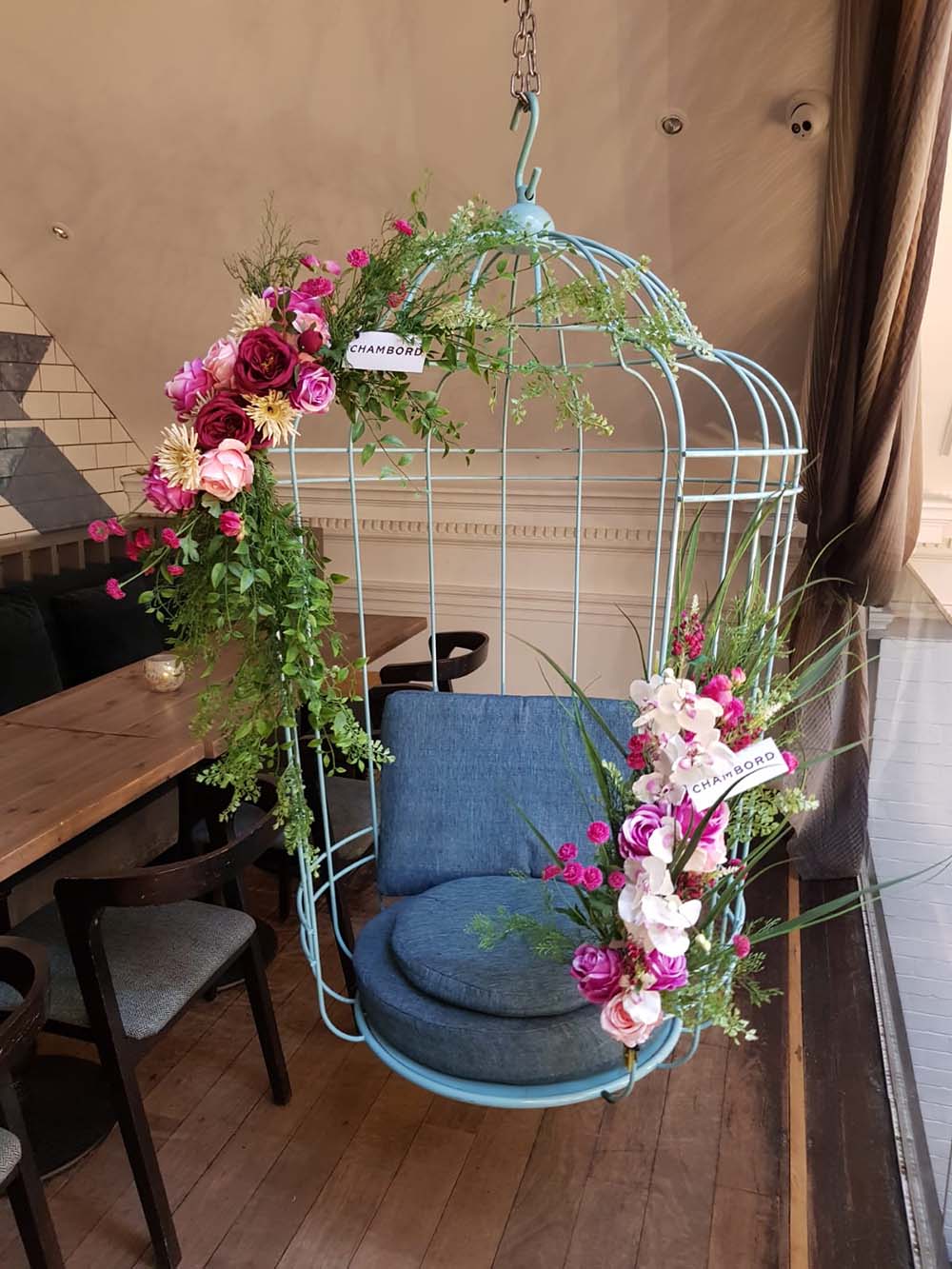 A hanging birdcage seat in Hummingbird Glasgow adorned with artificial floral displays and Chambord branding