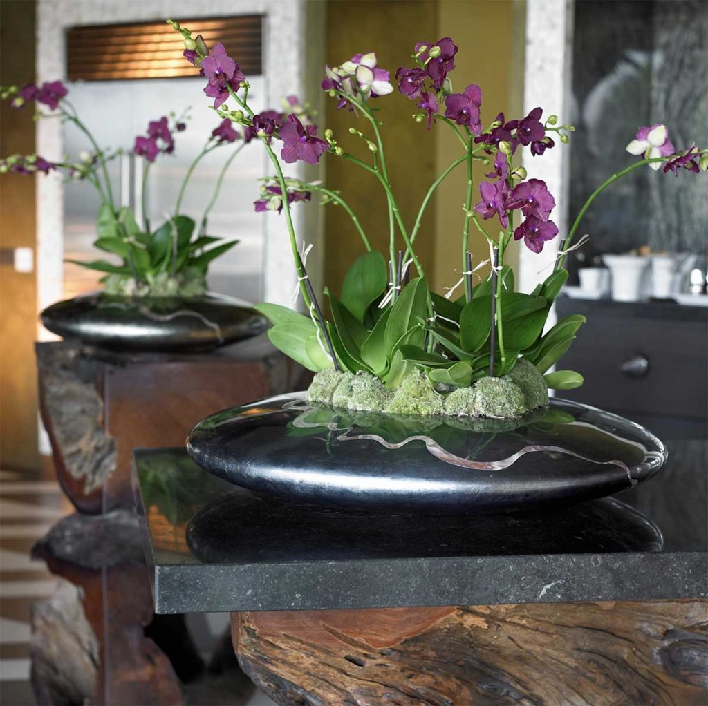 Purple orchids with a moss base in a stone effect circular planter atop a marble square sitting on a wooden bar