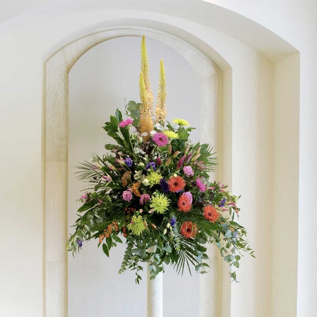 Fresh mixed flower pedestal arrangement with bright gerbera and leafy foliage in a hotel archway