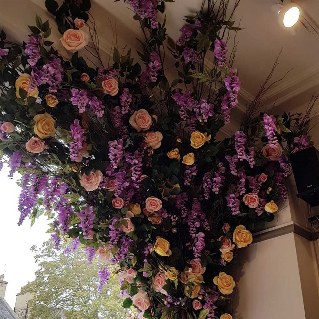 An explosion of colour for a window. Artificial yellow roses and purple wisteria draped over twisted willow