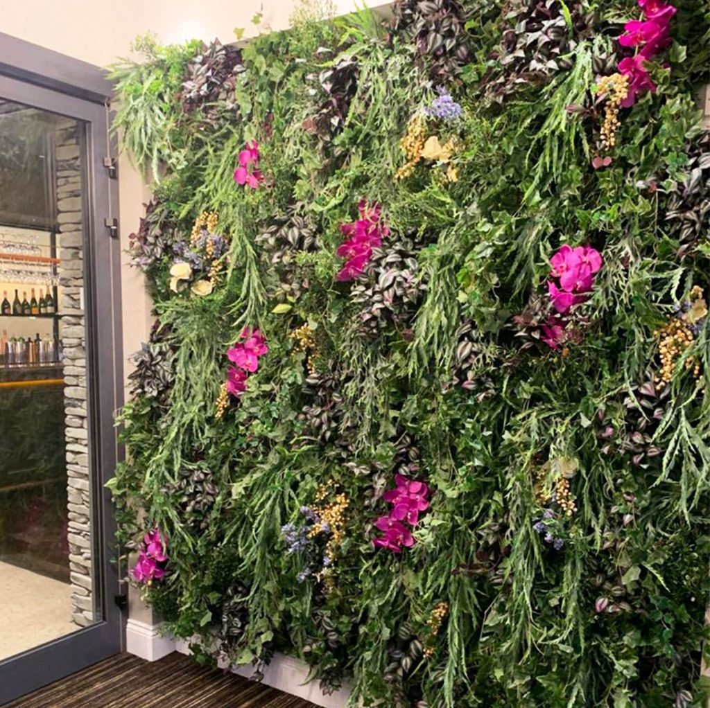 minimalist flower wall featuring mainly green foliage, with pink/ purple orchids and flowers interspersed within to create a winter floral wall display