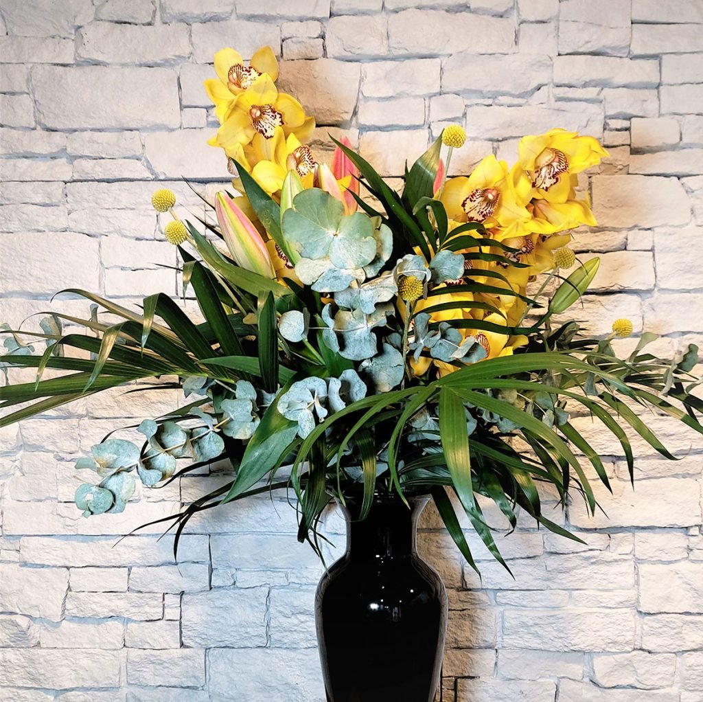 tall corporate flower display in black vase with yellow orchids, pink lilies, eucalyptus and palm leaves