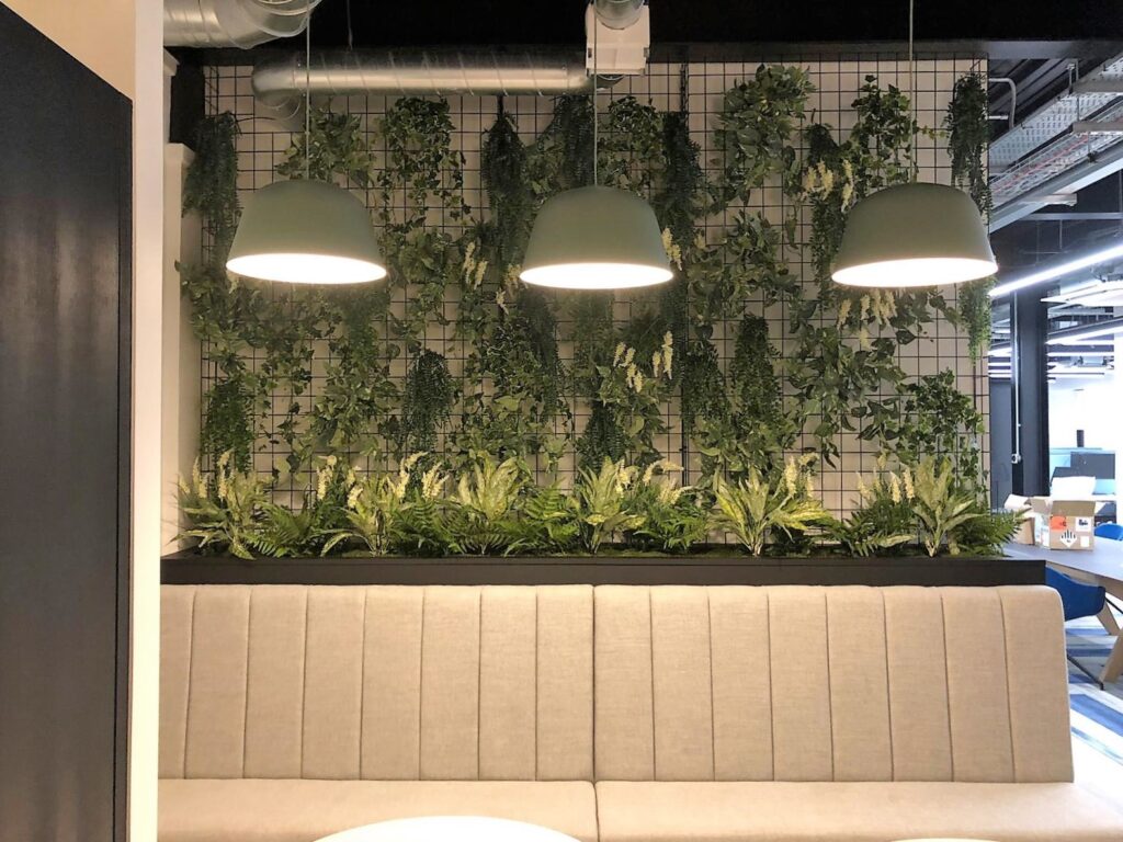 A mixture of artificial greenery secured to a mesh grid above a booth with trough planters and artificial plants.