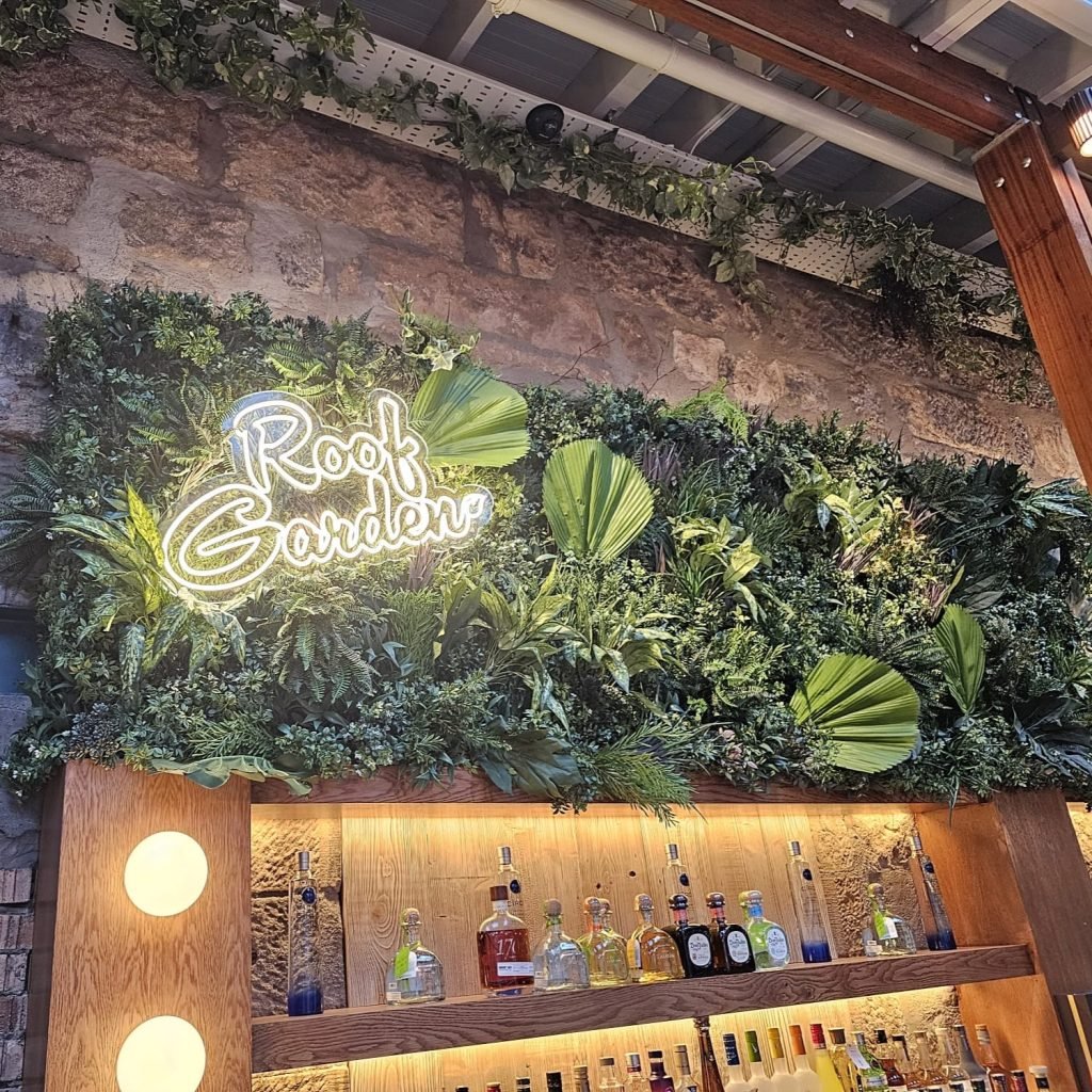 A decorative artificial green wall with neon signage. Trails of artifical ivy above the green wall adorn the ceiling at the kong roof garden in glasgow.